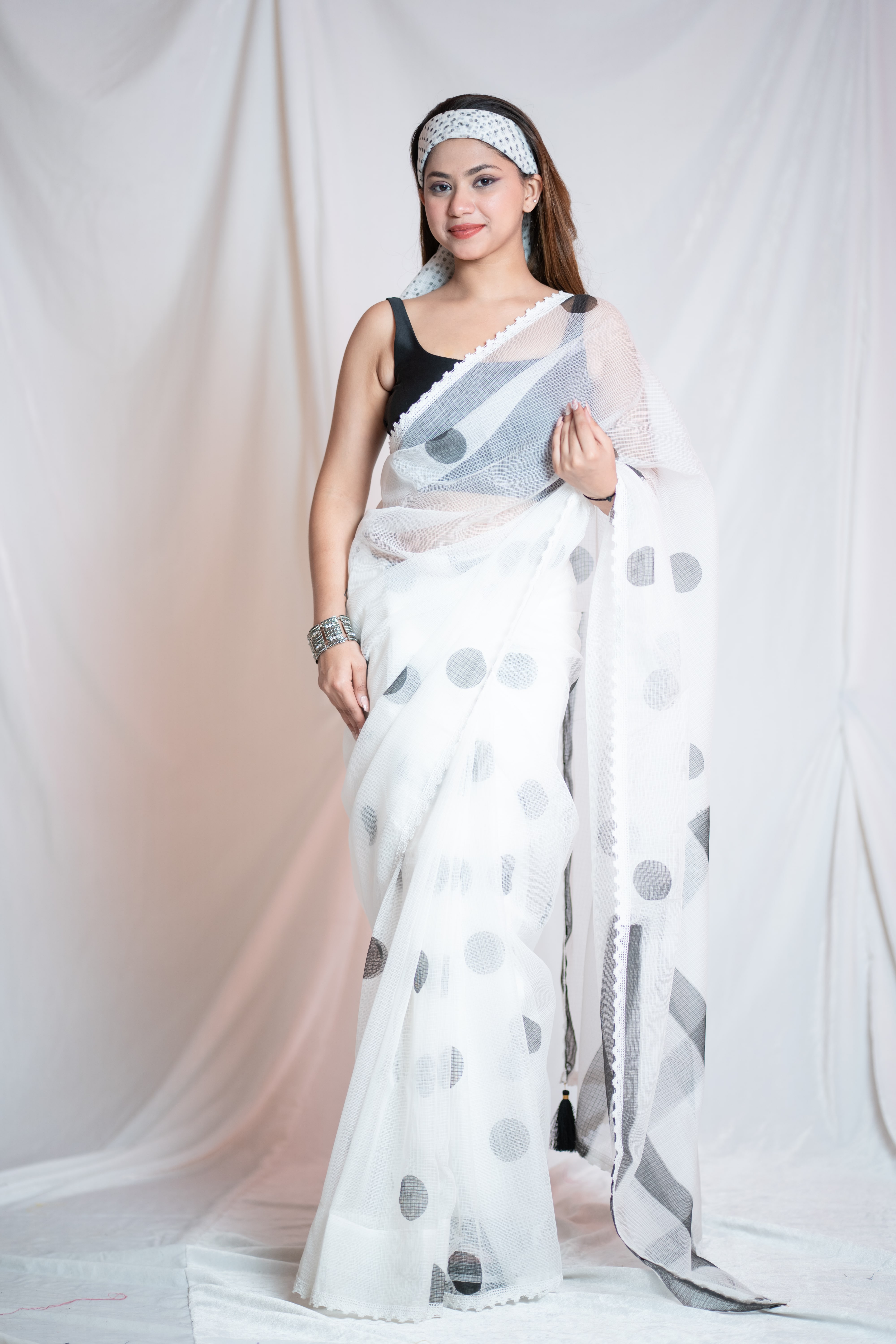White Kota Doria saree with black polka dots, featuring a lightweight, sheer fabric with a subtle sheen. The saree is draped elegantly, showcasing its fine texture and delicate pattern